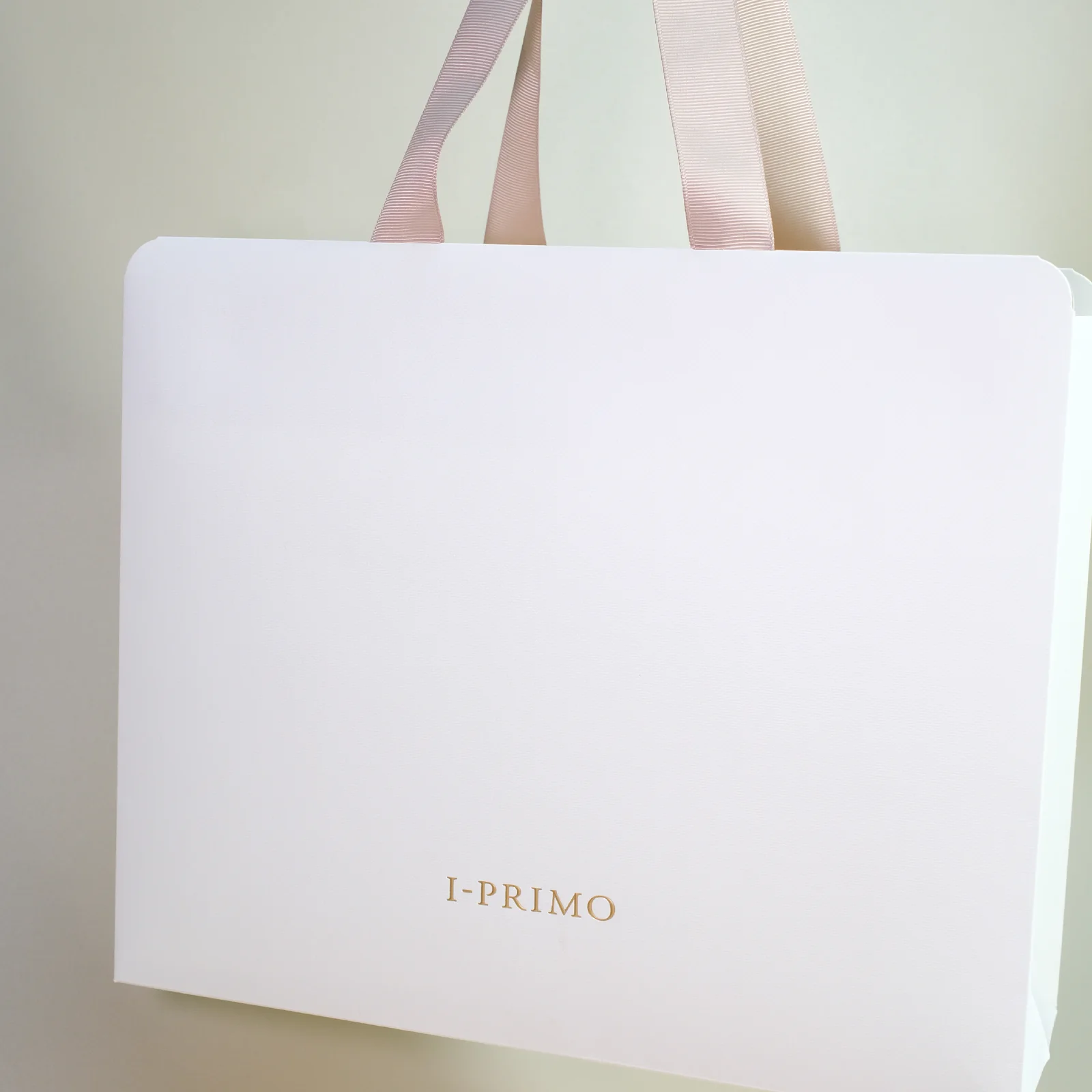 白色 I-PRIMO 紙袋，袋口圓角，粉色絲帶手挽，正面下方細小燙金 Logo